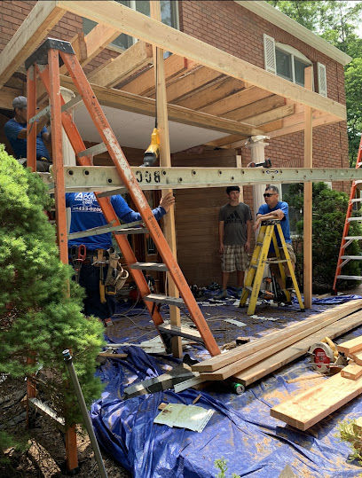Crew framing a covered porch addition beside a brick home, with ladders and lumber on site